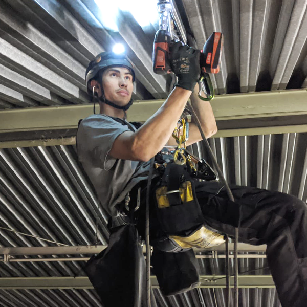Höhenarbeiter in Industriehalle mit Stirnlampe und Bohrmaschine unter Wellblechdach
